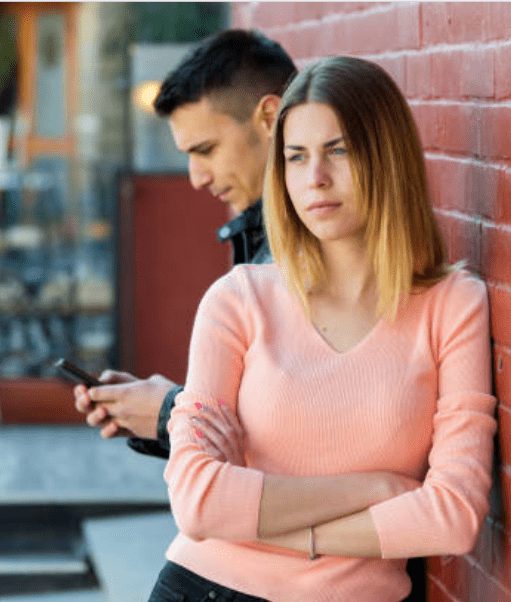 Nice Guy Syndrome: Are You Falling Into This Trap? 61 A Woman Leaning Against A Wall With Crossed Arms, Looking Distant, While A Man Stands In The Background Distracted On His Phone, Illustrating The Emotional Disconnect Nice Guys Often Face In Relationships.